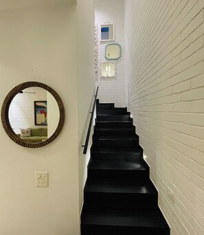 A staircase with white brick walls and black steps, leading to a landing with a framed picture and a light fixture.
