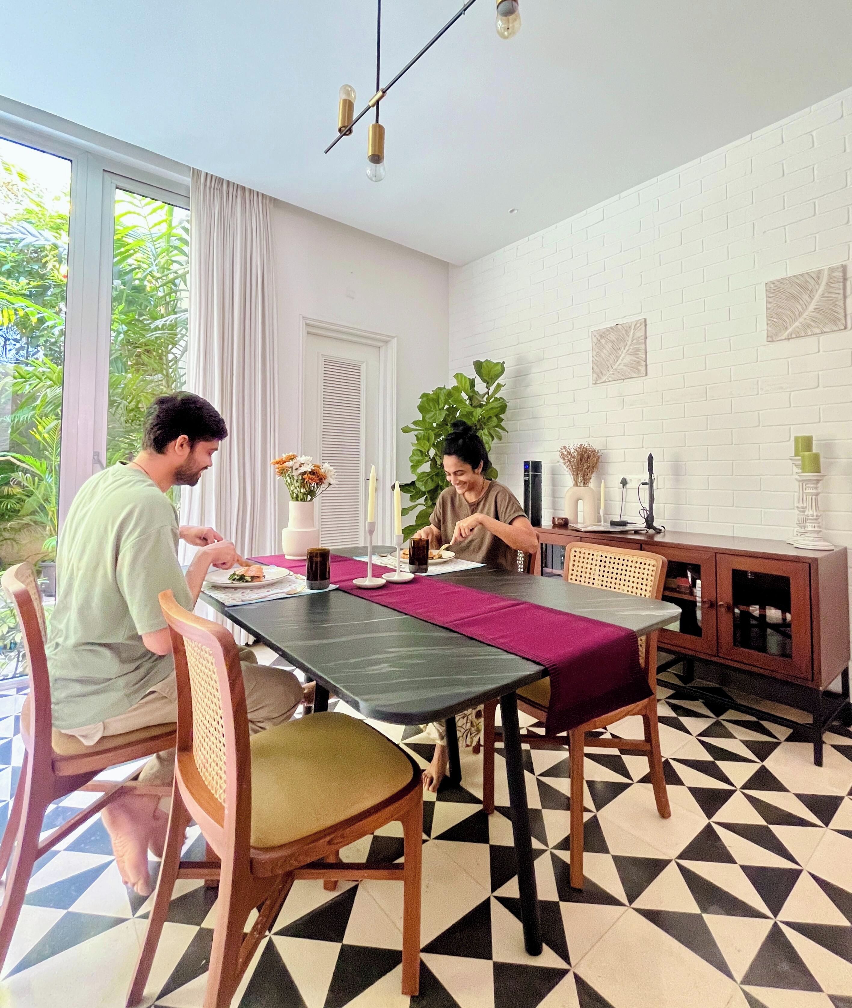 Un comedor con piso a cuadros blanco y negro, una mesa con mantel individual morado y una pared de ladrillo blanco con adornos de hojas.