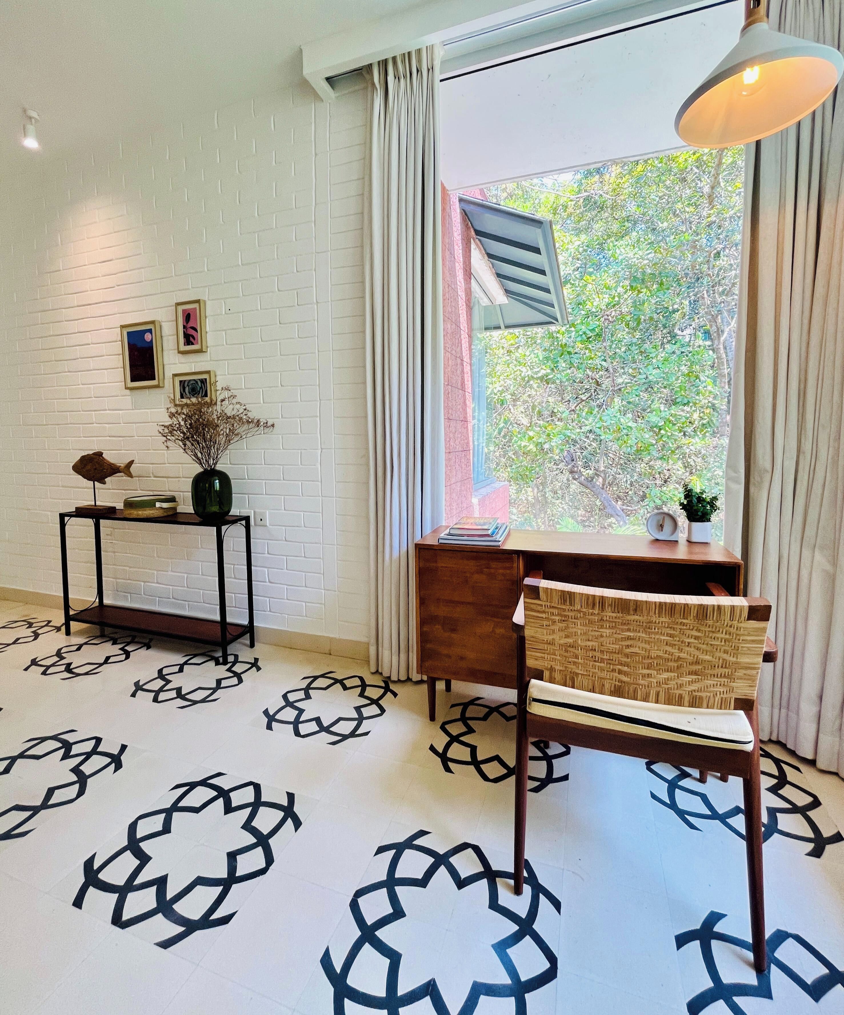 A room with a desk, chair, and a window with curtains. There is a wall-mounted shelf with decorative items and a patterned floor.