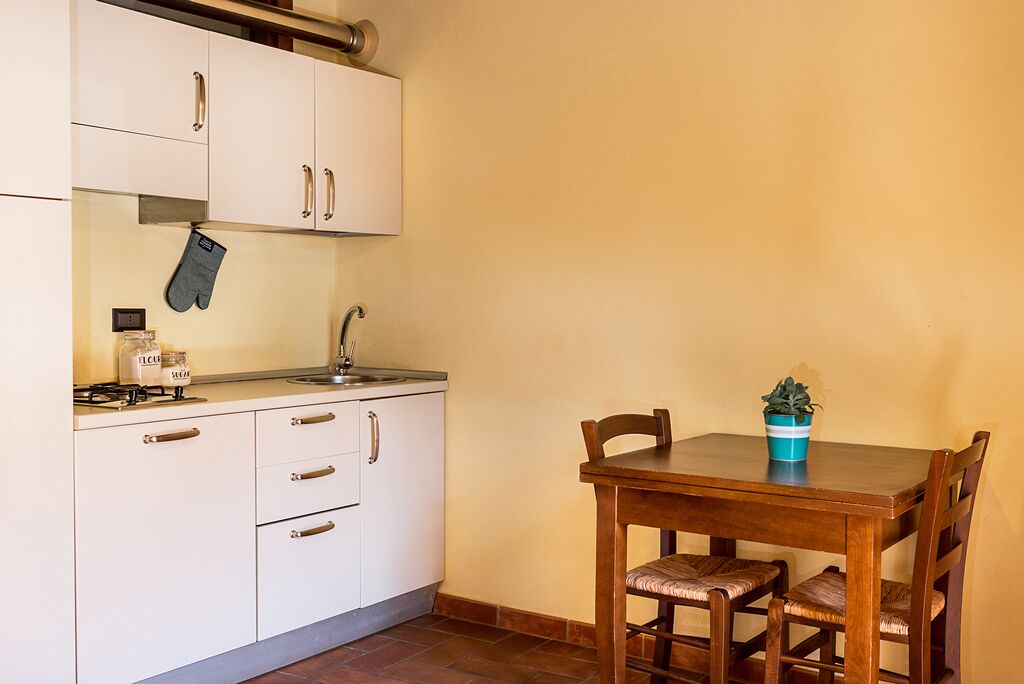 Una cocina con armarios blancos, una estufa y un lavamanos. Hay una mesa de madera con sillas y una planta en maceta.