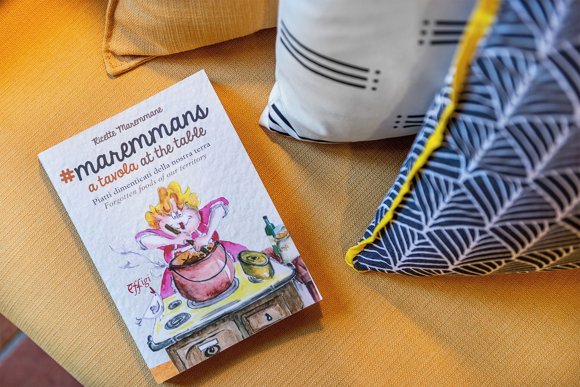 A book titled 'Ricette Marennane a tavola' with a kitchen illustration on a textured orange surface, next to a patterned pillow and a white bag.