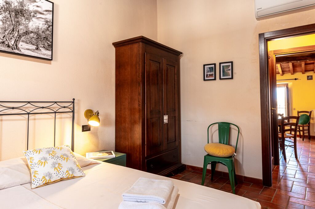 A hotel room with a bed, a wooden wardrobe, a green chair, and a wall-mounted lamp.