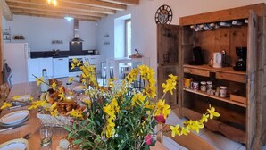 A modern kitchen with white cabinets, a wooden dining table set with yellow flowers, and a rustic wooden shelf with kitchen items. - Renovated country house, ideal for 10, natural setting and modern comfort (Gumières)