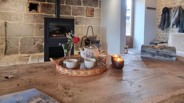 A rustic room with a stone wall, a black wood-burning stove, a wicker basket with cups and a teapot, and a candle on a wooden table. - Renovated country house, ideal for 10, natural setting and modern comfort (Gumières)