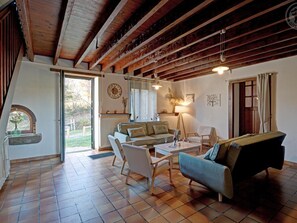 Ein geräumiges Wohnzimmer mit hohen Decken, Holzbalken, einer Couch, Sesseln, einem Couchtisch und Blick ins Freie.