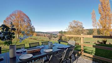 An outdoor dining area with a table set for four, surrounded by chairs, with a view of trees and hills in the background.