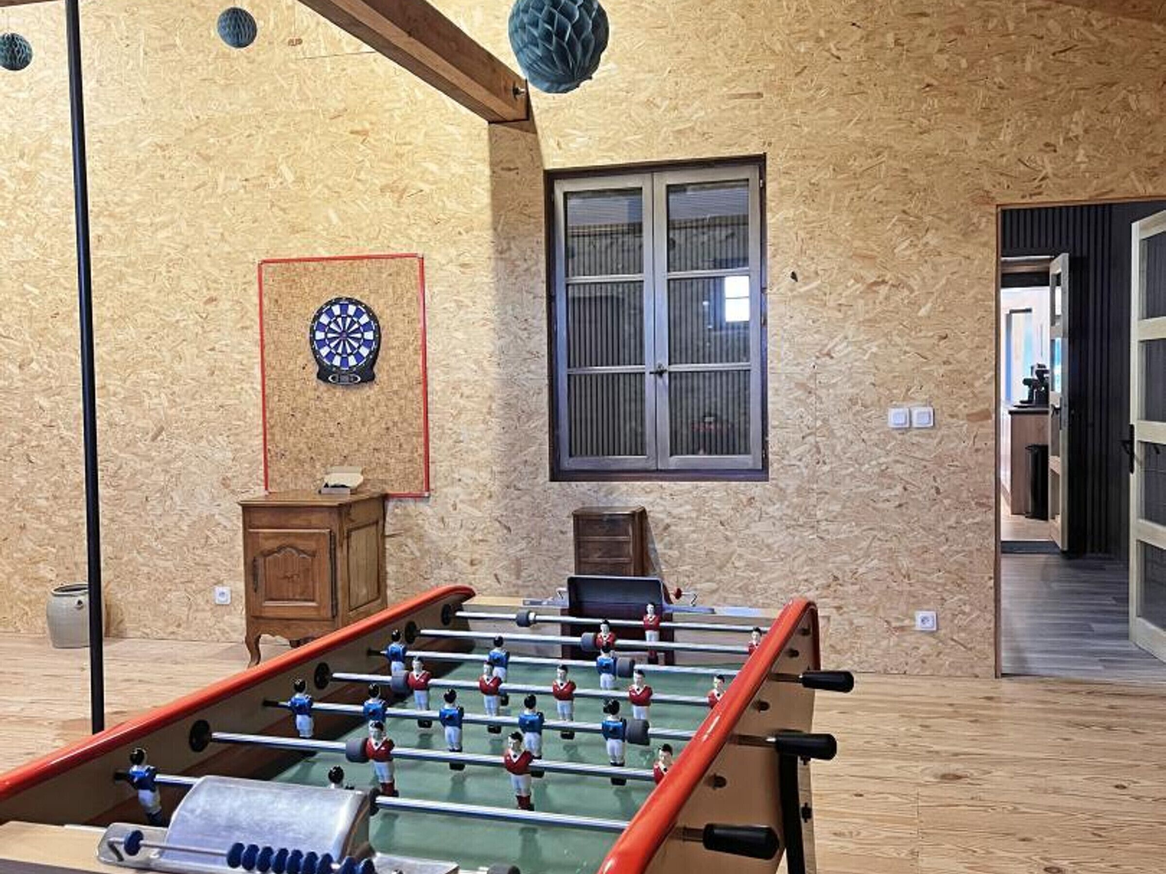 A room with a foosball table, a dartboard, and a window with blinds.