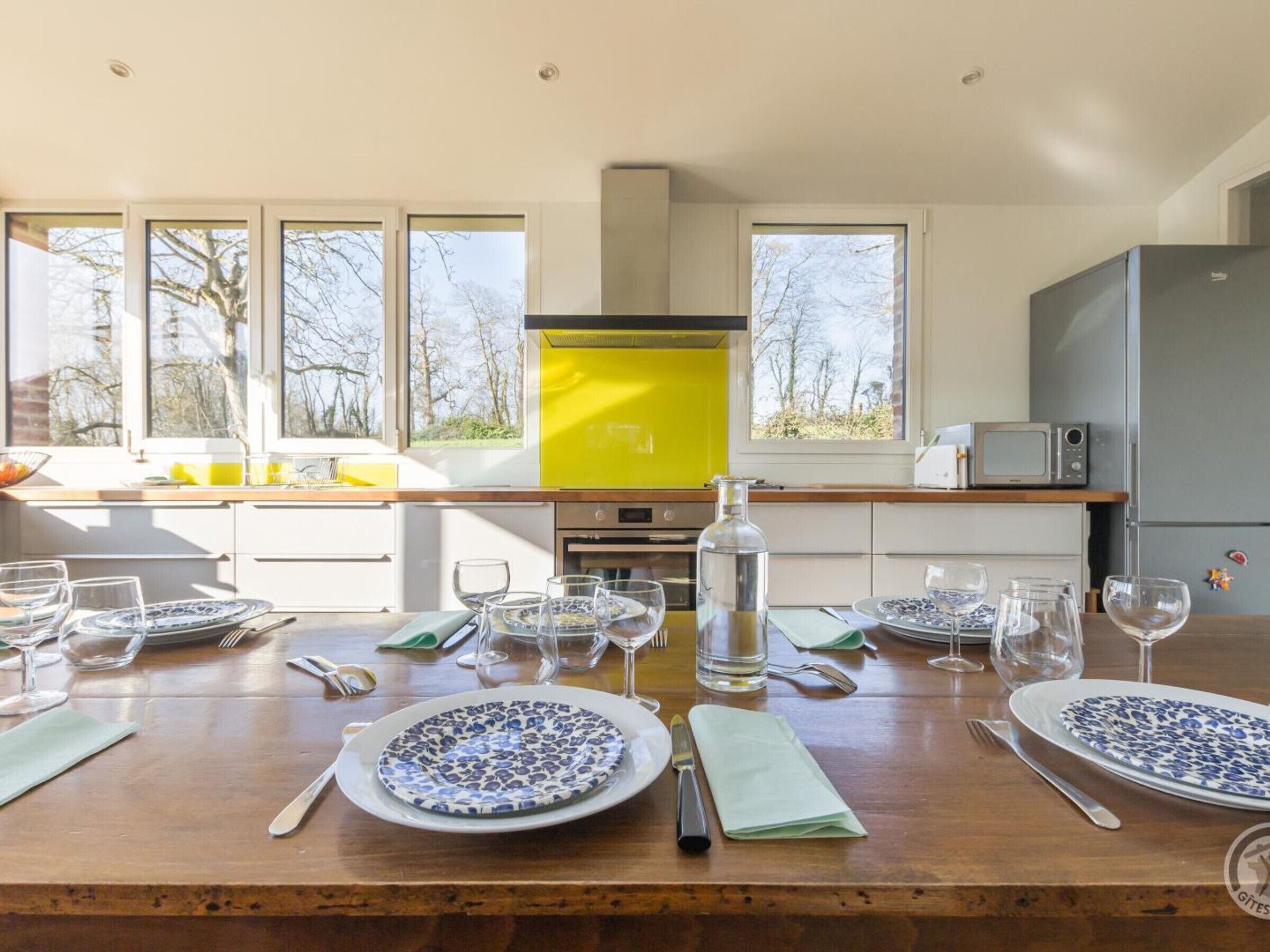 A modern kitchen with large windows, a yellow range hood, and a dining table set for four.