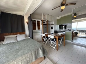 A bedroom with a bed, a dining area with a table and chairs, a kitchenette with a microwave and stove, and a view of the sea. - Le Grau d'Agde - Comfortable Studio for 4 People (Le Grau d'Agde)