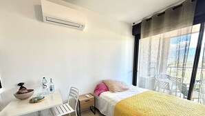 Habitación de hotel con cama, escritorio, silla, mesita, aire acondicionado, ventana con vistas y balcón.