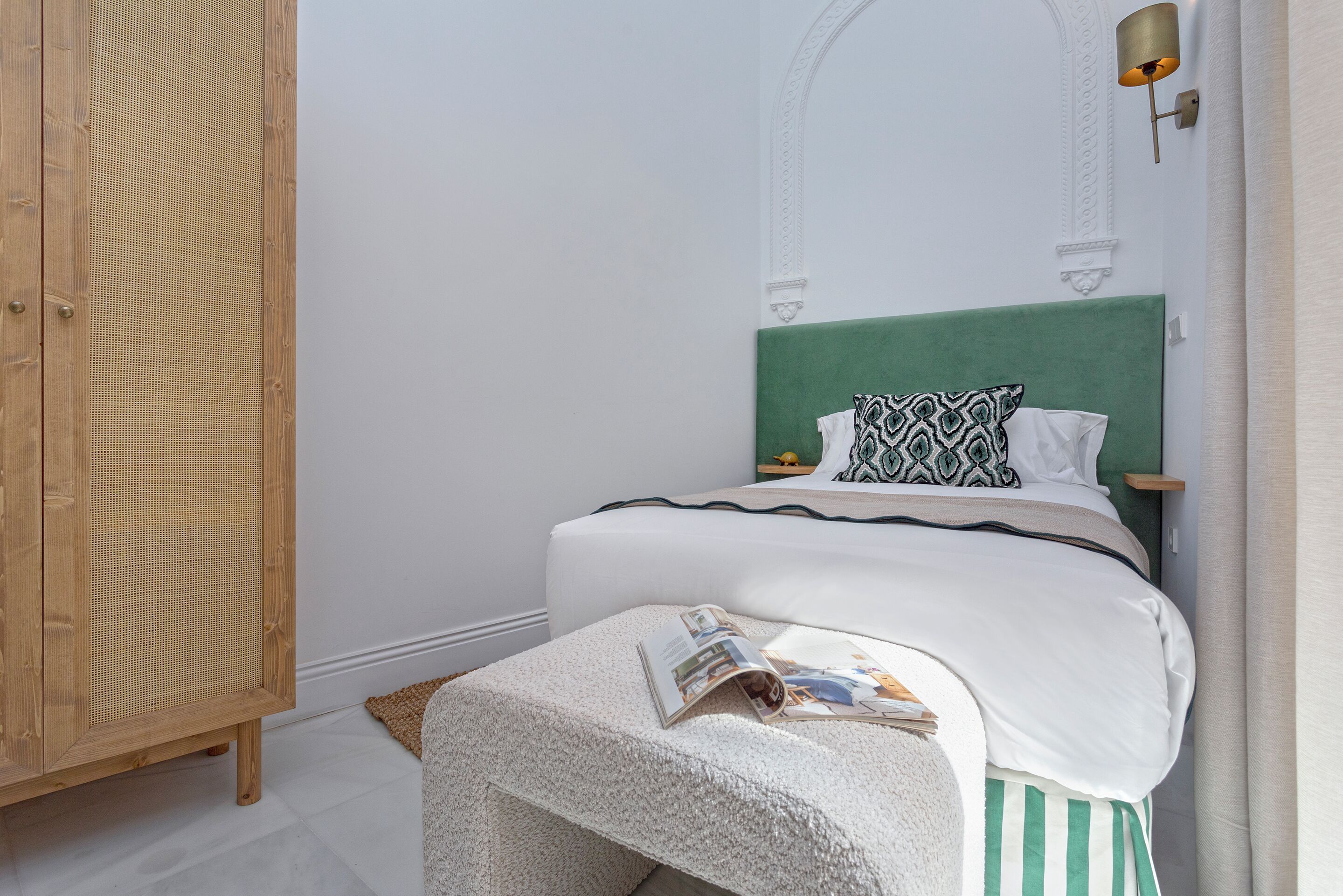 Une chambre à coucher bien rangée, avec une tête de lit verte, une table de chevet sur laquelle se trouve un magazine et une armoire en bois.