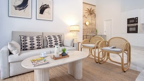 A modern living room with a sofa, coffee table, and wicker chairs. There are two framed abstract paintings on the wall, a lamp, and a kitchen area with an oven. - Cozy 3BD in Seville Center. Alfalfa (Seville)