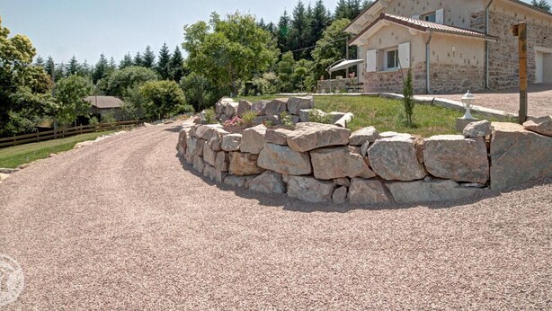 Una casa de piedra con camino de grava, rodeada de árboles y un muro de piedra.