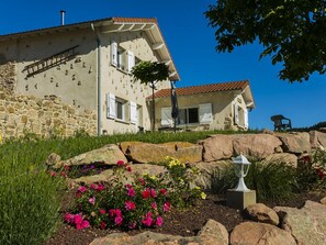 A two-story stone building with a sloped roof, white shutters, and a stone wall on one side.