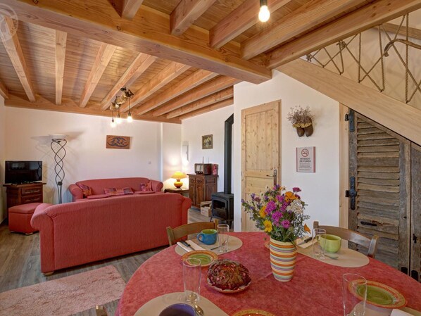 A cozy living room with a red sofa, a dining table with a vase of flowers, and a wooden ceiling.