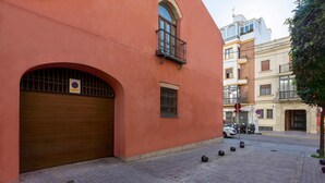 Un bâtiment avec un portail de garage fermé, une petite fenêtre avec un balcon, et une rue bordée de voitures garées et de bornes.
