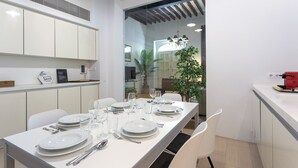Un espace de salle à manger moderne avec une longue table dressée pour quatre personnes, des chaises blanches et une cuisine visible à travers une cloison en verre.