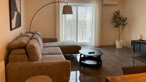 Ein modernes Wohnzimmer mit einer Eckcouch, einem runden Couchtisch, einer Topfpflanze und Blick durch ein großes Fenster.