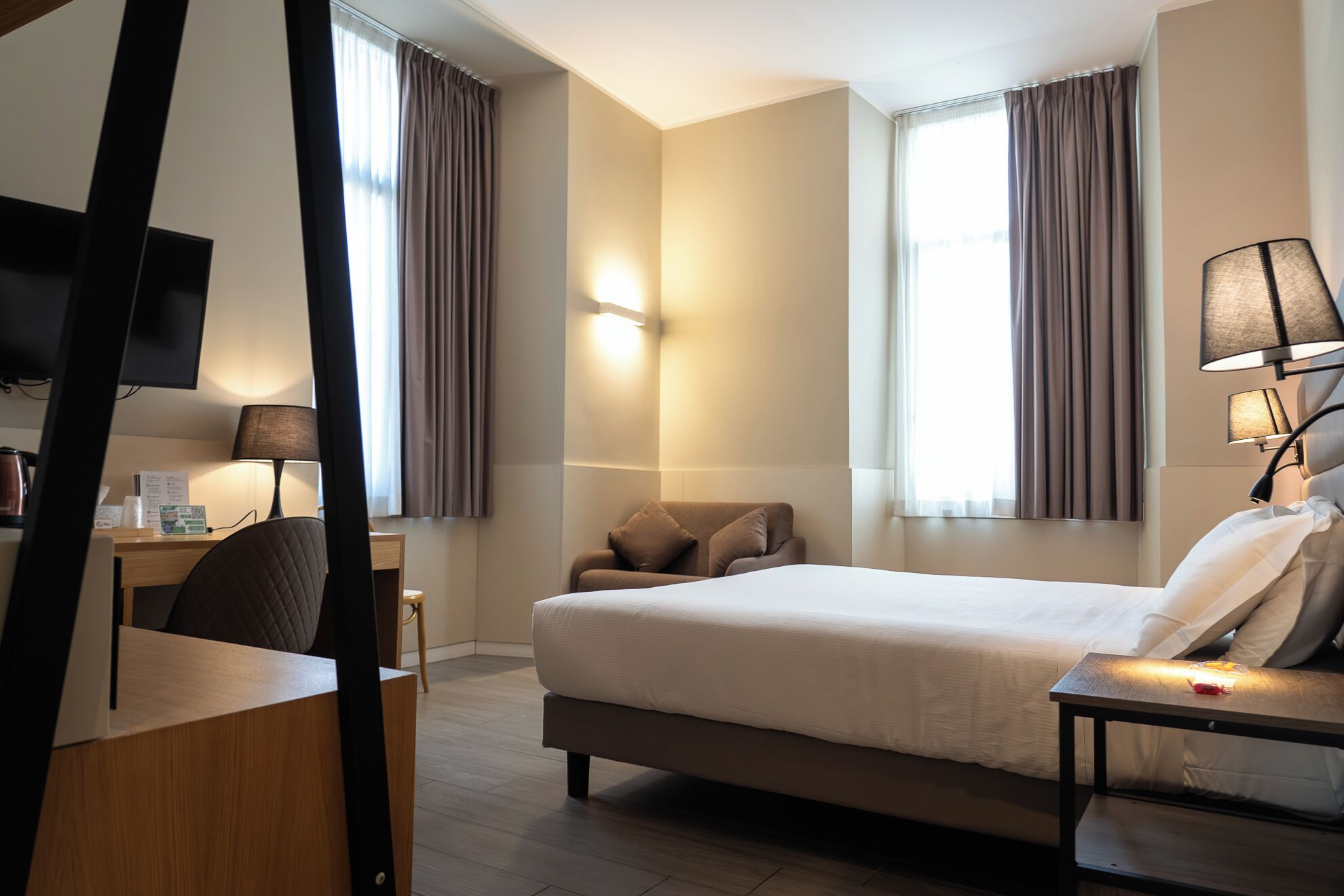 A hotel room with a bed, bedside table, lamp, chair, desk, TV, and curtains.