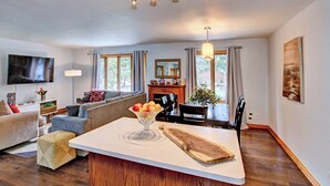 A modern living room with a sofa, a dining area with a table and chairs, a flat-screen TV mounted on the wall, and a fireplace.