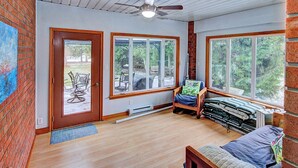 A room with a wooden floor, a bed, a window with a view of trees, a ceiling fan, and a door leading outside.