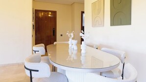 A modern dining area with a round glass table, white chairs, and minimalist decor. - Apartment Prestigia Agate - Golf view, Swimming pool & Comfort in Marrakech (Nîmes)