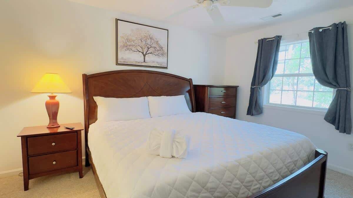 A neatly made bed with a quilted cover, a wooden nightstand with a lamp, a framed picture on the wall, and a window with curtains.
