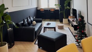 Un salon moderne avec un canapé noir, une petite table basse et un fauteuil jaune. On y trouve une fenêtre avec des rideaux, une plante en pot et un petit sapin de Noël.