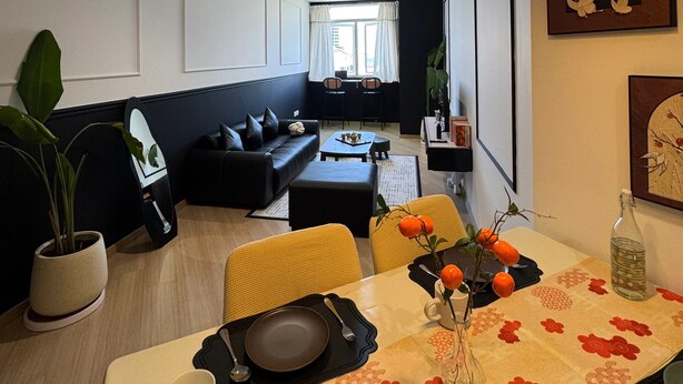 Un salon moderne avec un canapé noir, une table à manger décorée de fleurs orange et une grande fenêtre ornée de rideaux blancs.
