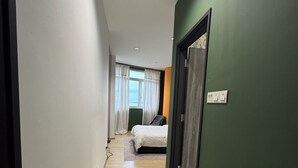 Un couloir étroit, doté d’un plancher en bois, mène à une pièce où se trouvent un lit et une fenêtre ornée de rideaux.