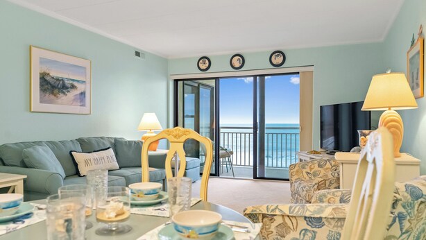 Sala de estar com vista para o oceano, mesa de jantar posta com pratos e copos, sofá e televisão.