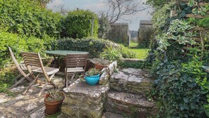 Un área de estar al aire libre con estilo rústico, con una mesa de piedra y sillas de madera, rodeada de abundante vegetación y un muro de piedra.