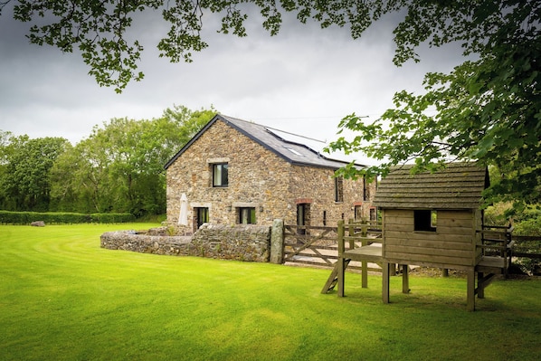 A stone house with a wooden outbuilding, surrounded by a well-maintained lawn and trees. - The Red Barn (Tavistock)
