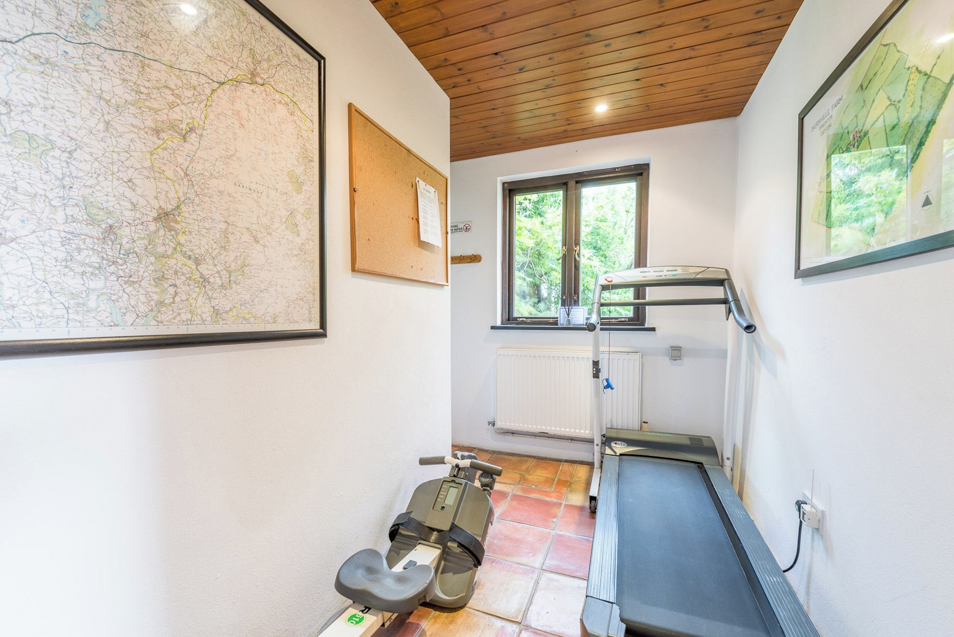 A small room with a treadmill, a map on the wall, and a window with a view of greenery.