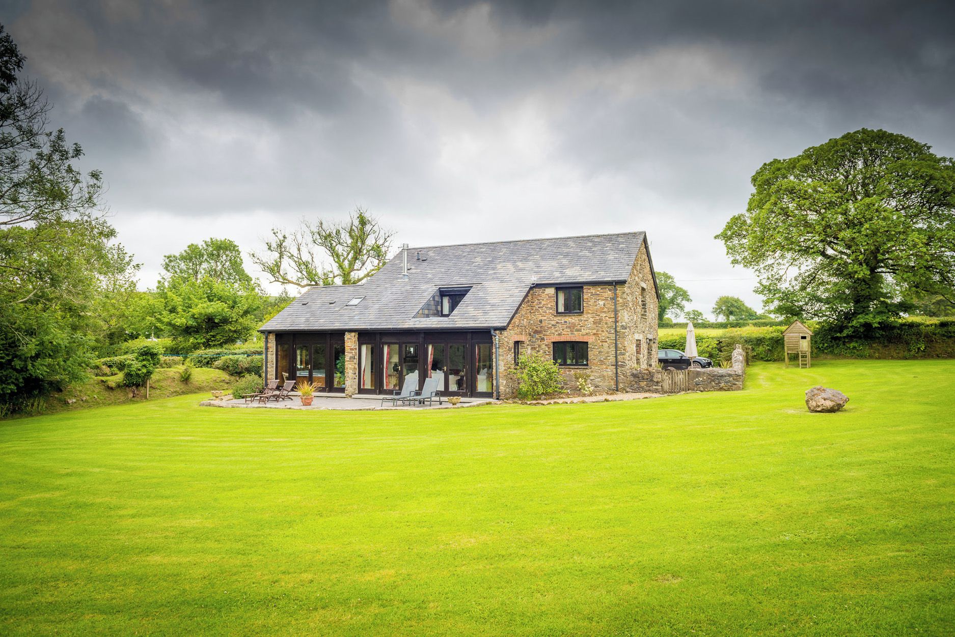 A stone house with a large lawn, a patio, and a car parked outside.