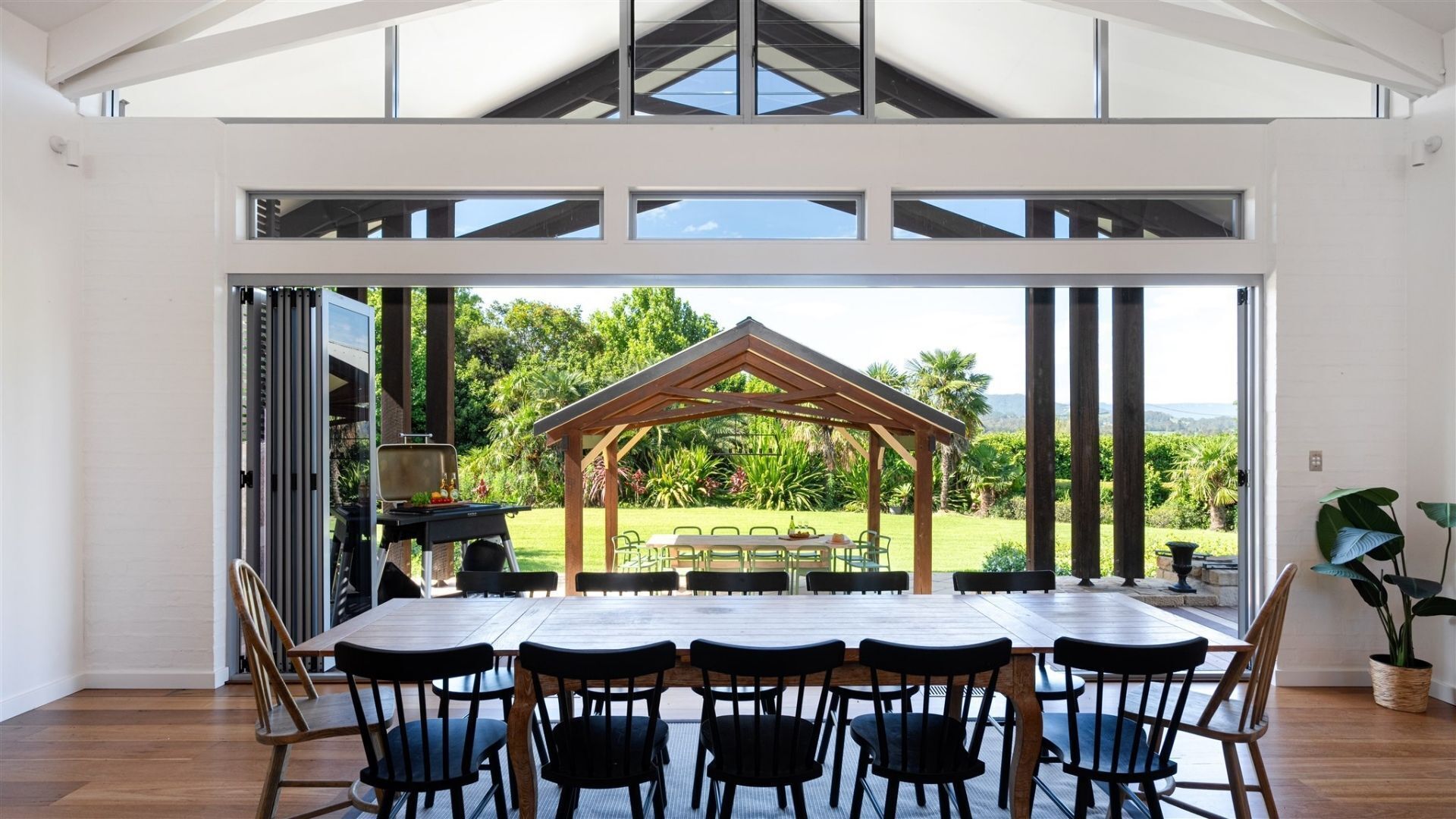 A spacious dining area with a large table and chairs, a wooden pergola, and a view of a lush garden and mountains.