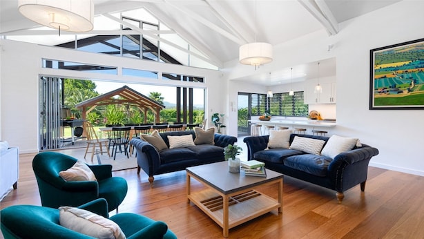 Un vaste espace de vie comprenant deux canapés bleu foncé, une table basse en bois et des fauteuils turquoise. Une grande fenêtre donne sur une terrasse extérieure aménagée avec un ensemble de mobilier de jardin.