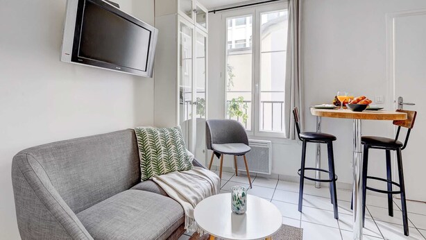 A modern living room with a grey sofa, a small round table, a TV mounted on the wall, and a dining area with a table and two chairs. - Charming Studio near Eiffel Tower for 2 (Paris)