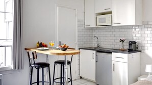 A compact kitchen with white cabinets, a microwave, and a small countertop. There is a wooden table with two stools, a fruit bowl, and a glass of orange juice. - Charming Studio near Eiffel Tower for 2 (Paris)