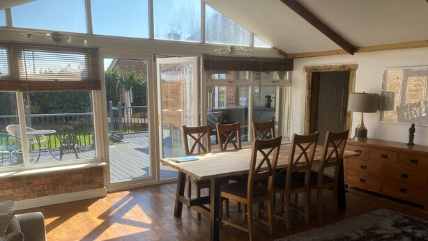 Ein geräumiges Zimmer mit einem Holzessttisch und Stühlen, einer Couch, einem Holzschrank und großen Fenstern mit Blick auf eine Terrasse und Gartenmöbel.