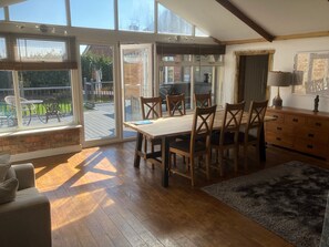 A spacious room with a wooden dining table and chairs, a sofa, a wooden cabinet, and large windows offering views of a deck and outdoor furniture. - Wall Heath Retreat - Period 3 Bedroom Cottage (Kingswinford)
