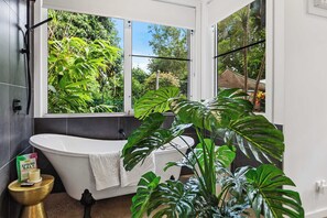 Un bagno moderno con una vasca freestanding, una finestra vista su vegetazione rigogliosa e un piccolo tavolino con articoli da toilette.