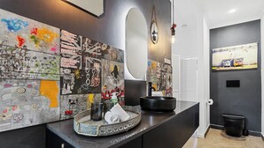 A modern bathroom with a black vanity, a large mirror, and a wall adorned with colorful graffiti art. - Tropical Family Retreat with Pool and Garden (Machans Beach)