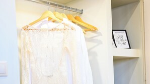 A closet with a white long-sleeve top hanging on a wooden hanger, a black speaker on the floor, and shelves with baskets containing folded clothes. - Cozy Studio 2 (Malia)