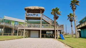 A two-story building with a thatched-roof structure on the upper level, a covered outdoor area, and a slide. - La Palapa whole house at pedestrian beach! Surfside Beach Texas! (Freeport)