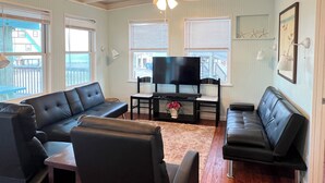 A living room with a black leather sofa, a TV on a stand, and a ceiling fan. - La Palapa whole house at pedestrian beach! Surfside Beach Texas! (Freeport)