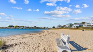 A sandy beach with a white bench, a clear blue sky, and houses in the background. - Cape Cod Bungalow Walk to Beach Firepit (Yarmouth)