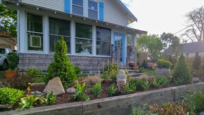 A two-story house with a front porch, a small garden with various plants and rocks, and a clear sky. - Cape Cod Bungalow Walk to Beach Firepit (Yarmouth)