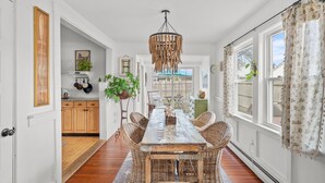 A dining area with a rustic wooden table and wicker chairs, a hanging light fixture, and a view to the outside. - Cape Cod Bungalow Walk to Beach Firepit (Yarmouth)