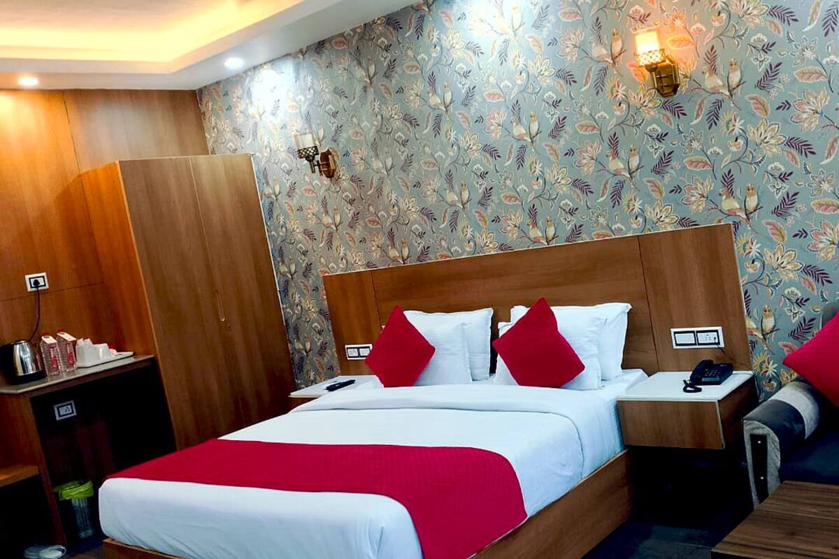 A hotel room with a wooden bed, white bedding, red pillows, a wooden nightstand, a wooden wardrobe, and a floral wallpaper.